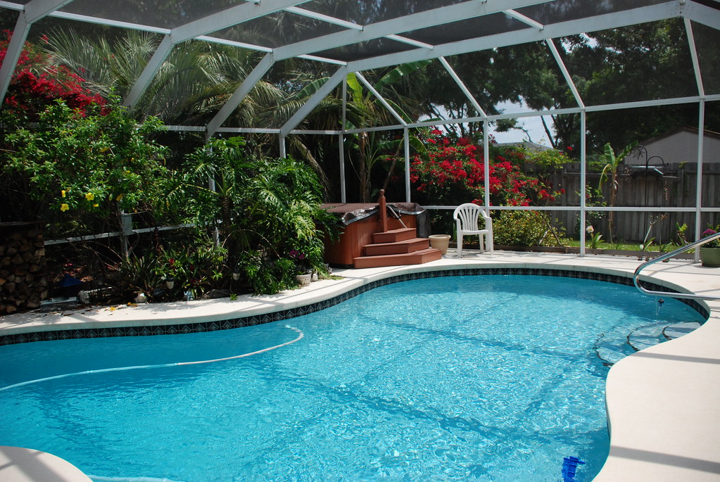 White-framed pool screen enclosure over a kidney-shaped pool with a hot tub and tropical planting