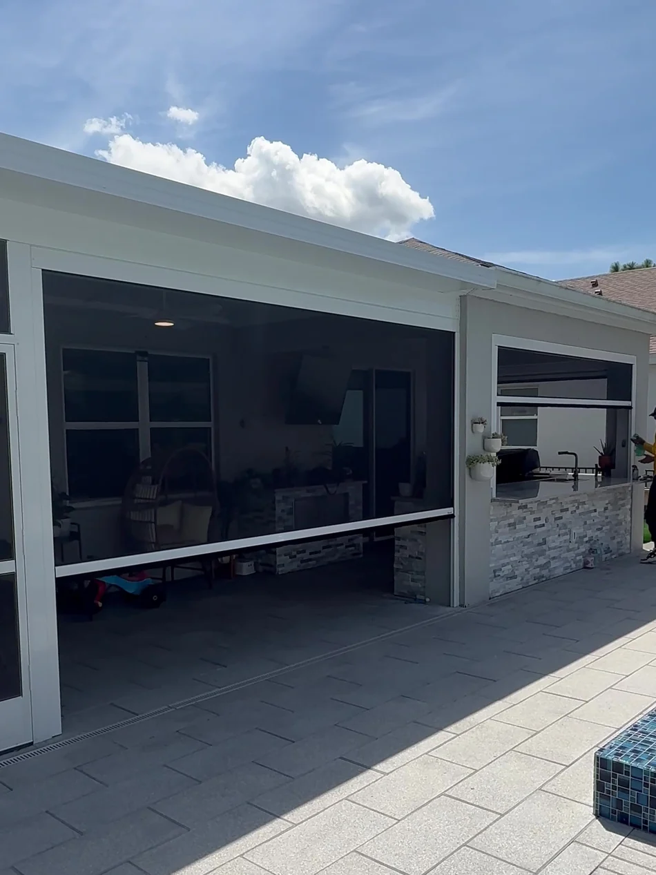 Motorized retractable screen half lowered over a lanai opening, showing the aluminum housing above and the outdoor kitchen behind