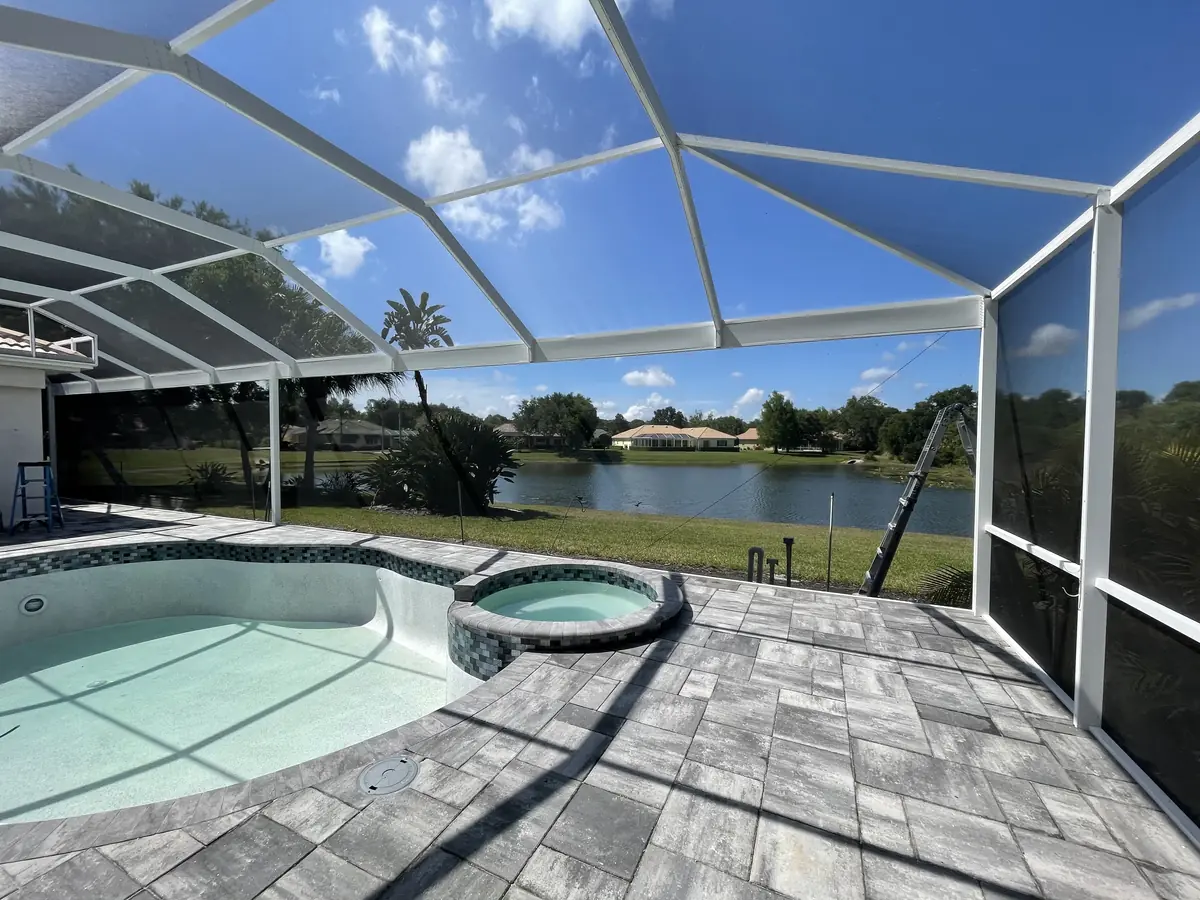 White aluminum pool cage over a paver pool deck and spa with a lake behind it